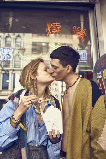 A couple shares a kiss in front of a restaurant with glowing neon signs, while one person holds a take-out container of noodles and chopsticks.