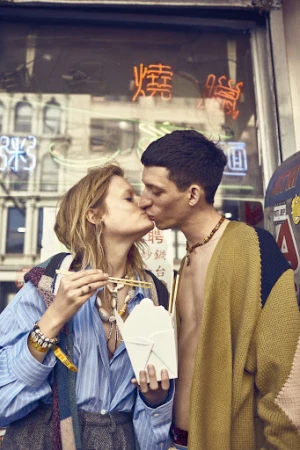A couple shares a kiss in front of a restaurant with glowing neon signs, while one person holds a take-out container of noodles and chopsticks.