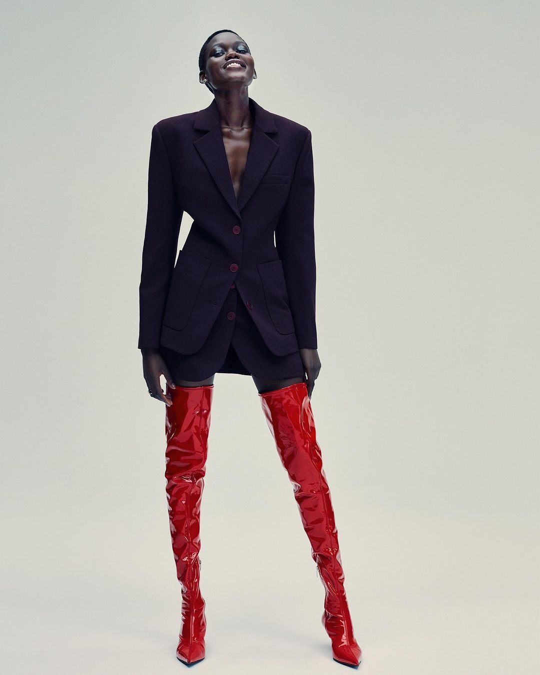 Stylish black model poses in a short black skirt suit and red patent leather thigh-high boots against a plain backdrop in a studio shot.