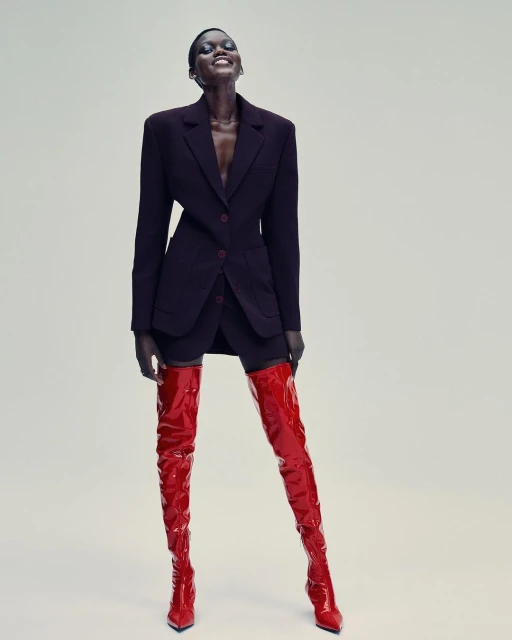 Stylish black model poses in a short black skirt suit and red patent leather thigh-high boots against a plain backdrop in a studio shot.