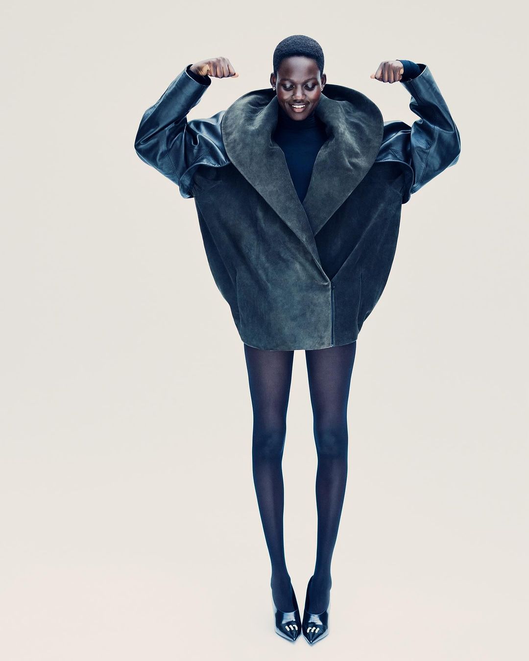 Fashion model in oversized dark green suede jacket, black tights, and pointed black heels poses on a pale beige background.