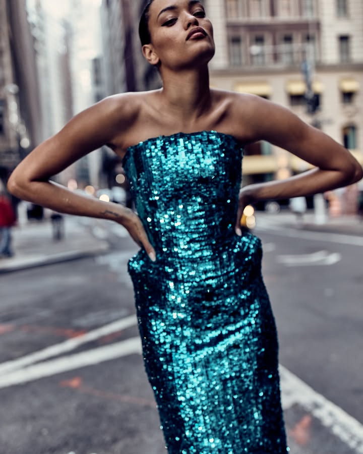 Woman in a turquoise sequined gown poses in a city street with tall buildings in the background, hands on hips, looking upwards.