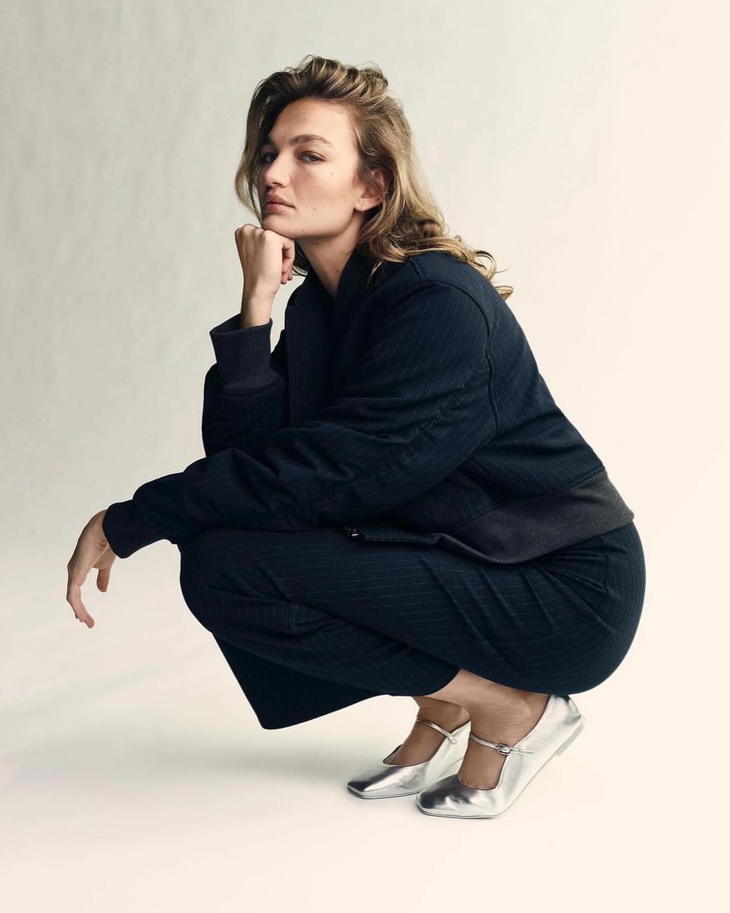 Blonde woman in navy bomber jacket and pinstripe pants poses in a studio with silver Mary Jane shoes, in a medium shot on white background.