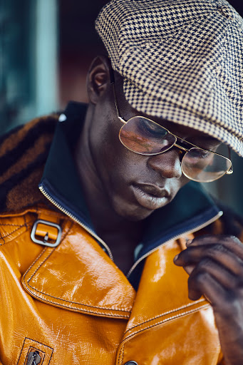 Stylish adult male poses in a yellow-gold jacket with a houndstooth hat and glasses in a close-up fashion portrait.