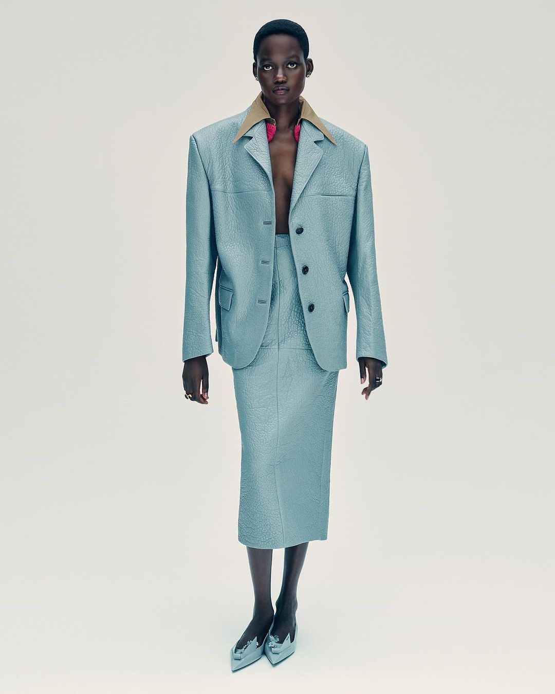 Dark-skinned fashion model poses in a textured light blue blazer and pencil skirt with metallic blue heels against a light background.