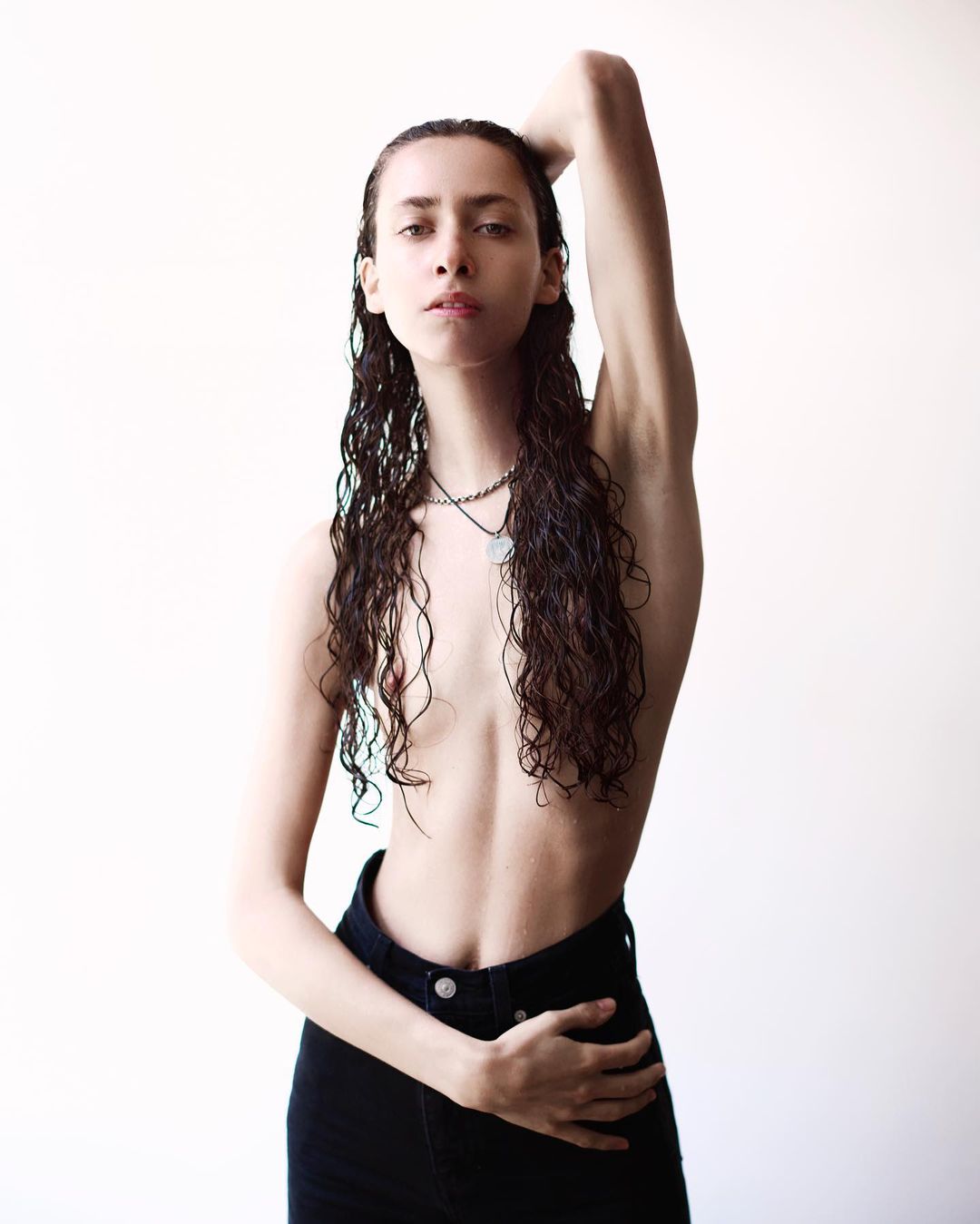 A young adult female with long dark curly hair stands in a photo studio, wearing jeans and a necklace, with a light neutral background.