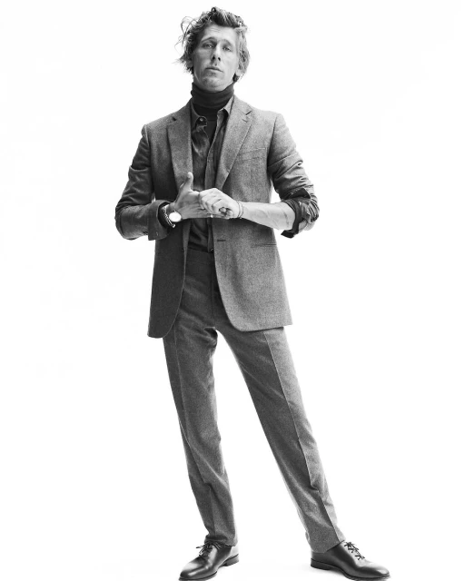 Full-length portrait of an adult male wearing a grey suit with a black turtleneck, standing against a white backdrop in a studio setting.