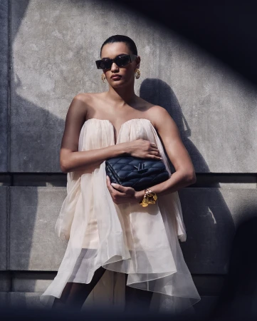 Model in strapless tulle dress and sunglasses holding a black quilted handbag against stone wall by Billy Kidd