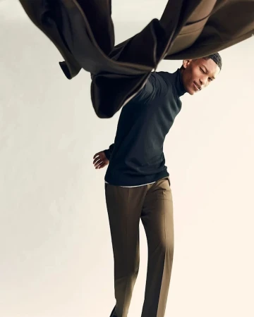 Young man in a dark turtleneck holding up a brown coat against a clean white seamless studio background.