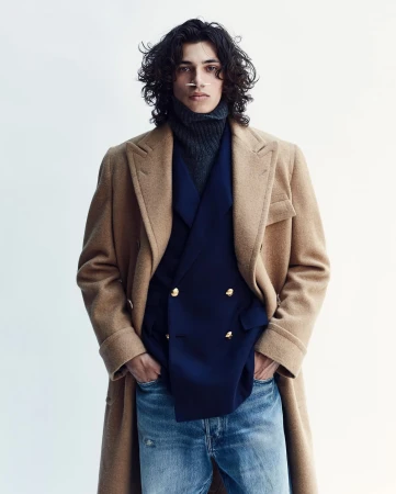 A man with long dark curly hair wearing a camel coat, navy blazer, grey turtleneck, and blue jeans against a light background.