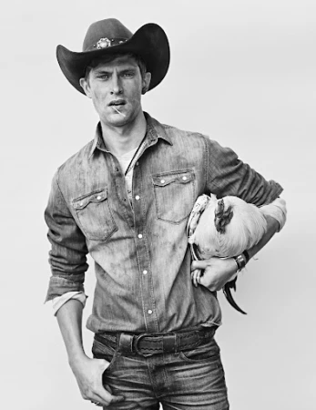 Monochrome portrait of a young man wearing a cowboy hat and denim shirt holding a rooster under his arm.