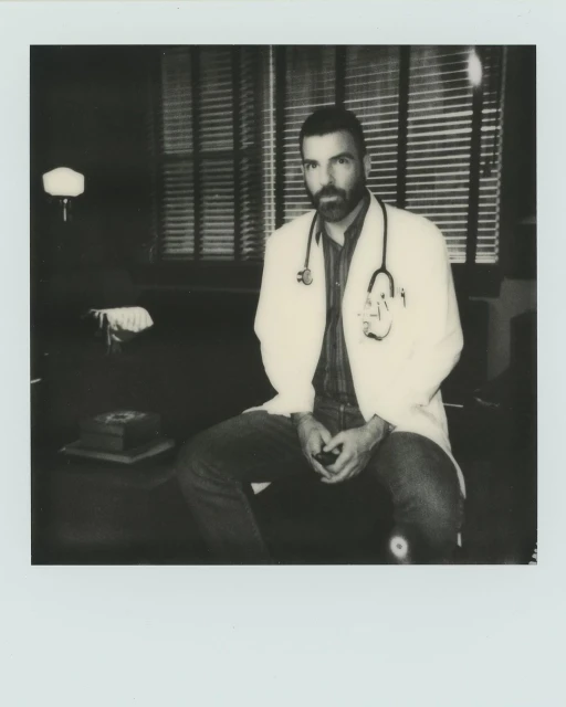Monochrome shot of a seated doctor wearing a white coat and stethoscope in an office setting with window blinds in the background.