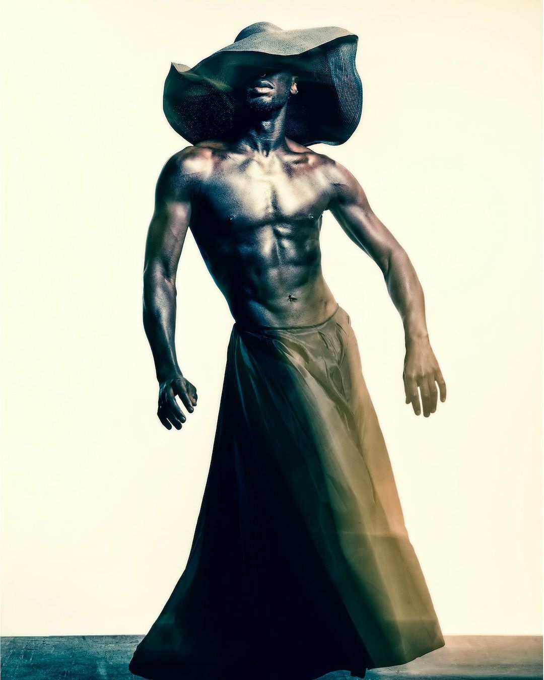Striking portrait of a Black man wearing a large hat and skirt-like garment, showcasing his physique in front of a plain backdrop.