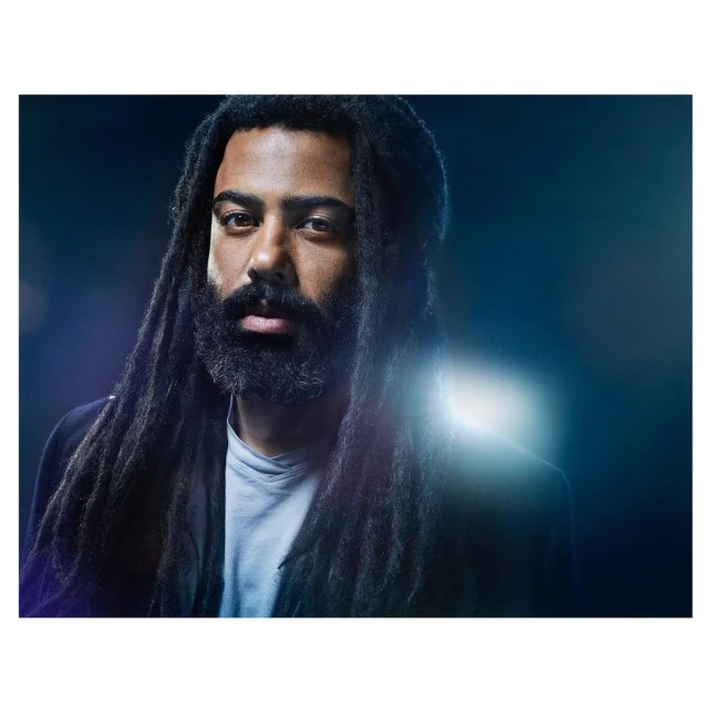 Portrait of an adult male with dark hair in dreadlocks and a beard wearing a blazer, in front of a dark blue background.