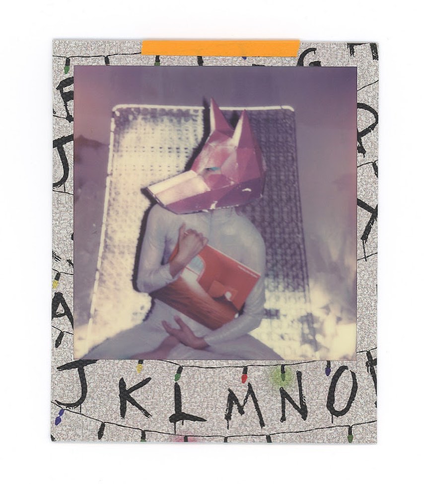 Person in a full bodysuit and a geometric fox head poses holding a red book in a photograph taped to a letter-covered backdrop.