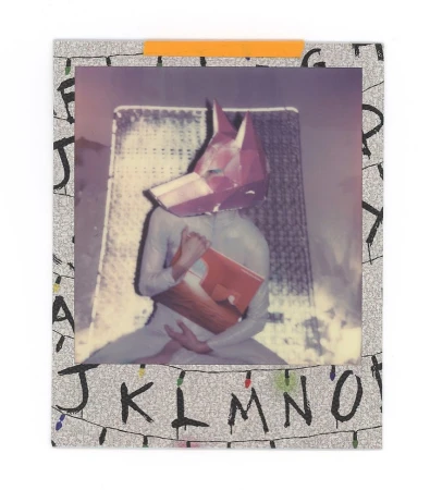 Person in a full bodysuit and a geometric fox head poses holding a red book in a photograph taped to a letter-covered backdrop.