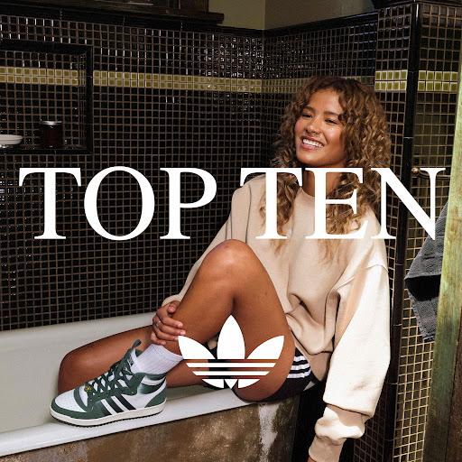 A woman with curly hair poses while sitting on the edge of a bathtub wearing sneakers with the Adidas logo displayed.