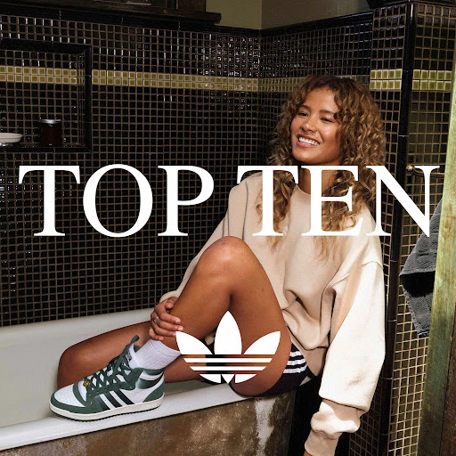A woman with curly hair poses while sitting on the edge of a bathtub wearing sneakers with the Adidas logo displayed.