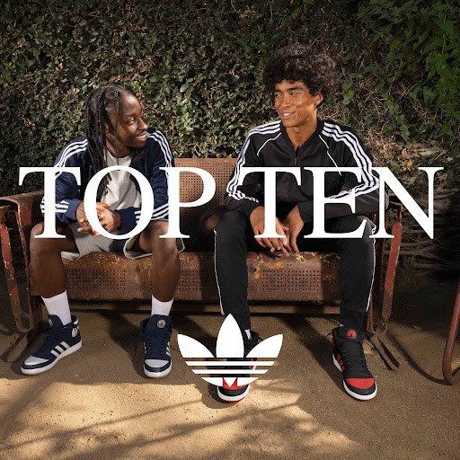Two men sit on a rusty bench in an outdoor setting, wearing athletic tracksuits and sneakers. Adidas logo and brand text are visible.