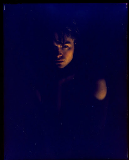 Low-key portrait of a young adult with fair skin in a dark, possibly indoor environment, lit by dim, warm-toned light.
