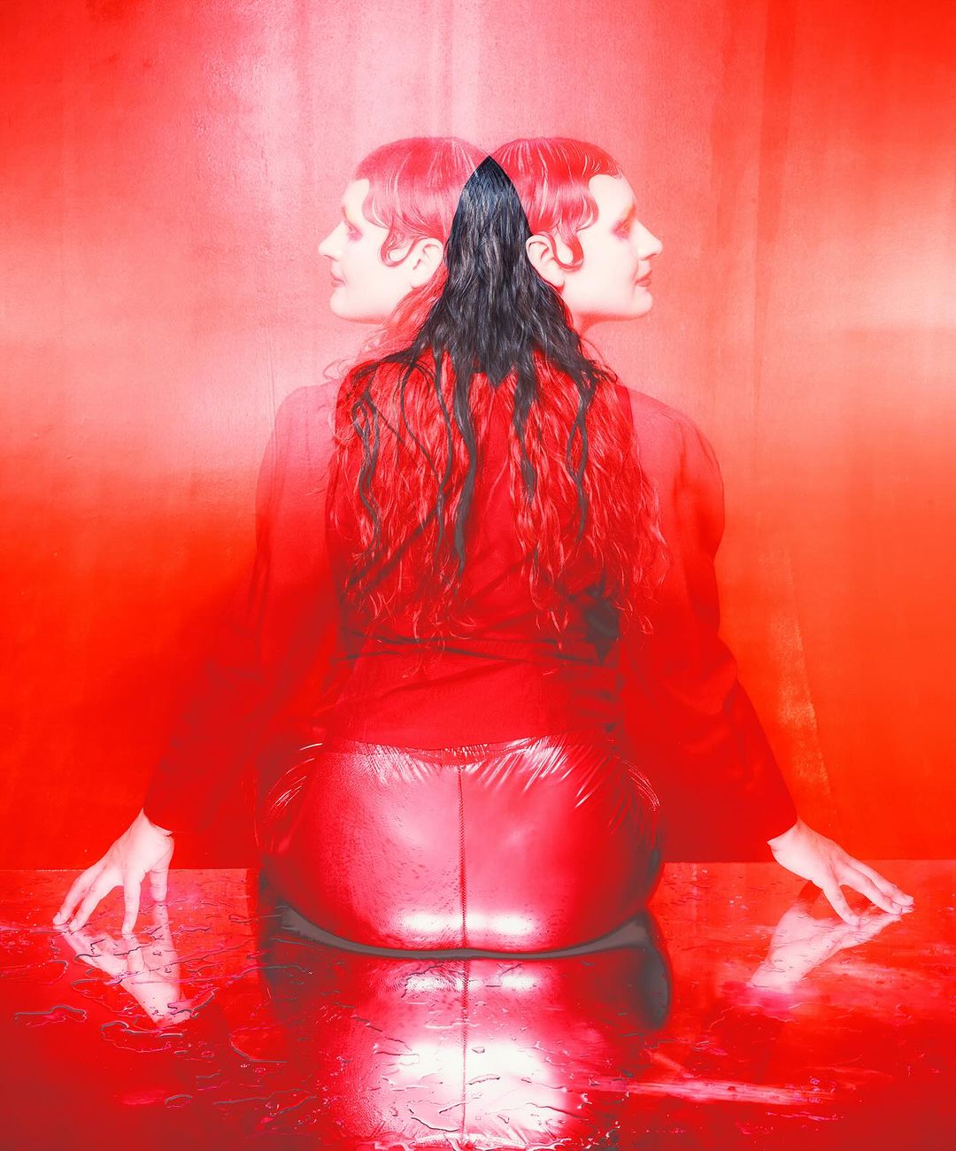 Two adult women with long hair are positioned back to back in a photo studio setting, bathed in monochrome red light, with reflections visible.