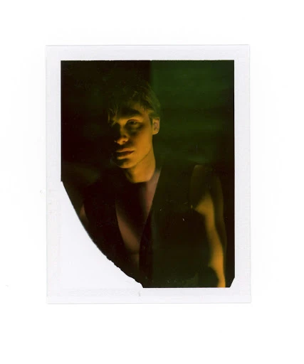 Polaroid photo of a young adult male with short blonde hair and a serious expression, wearing a dark top and posing indoors.