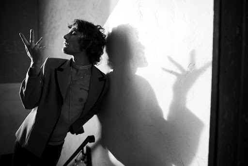 Monochrome portrait of a man leaning against a textured wall with a dramatic hand shadow cast across the surface.