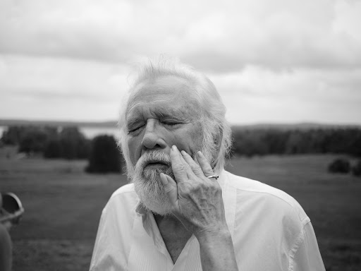 Monochrome portrait of an elderly man with white hair and beard, eyes closed, hand to face, in an outdoor field with a cloudy sky.