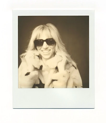Sepia portrait of a smiling woman with blond hair, sunglasses, and a fur coat captured in a vintage Polaroid instant photograph.