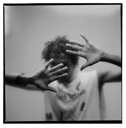 Portrait of an adult with dreadlocks, facing the camera with hands up, in front of a gray wall, with a square black frame.