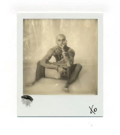 Polaroid-style photo of a bald man sitting and leaning on a vintage television, showcasing tattoos and jewelry, sepia-toned.