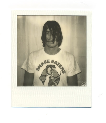 Indoor portrait of a young adult man wearing a t-shirt with 'Snake Eaters' logo in a Polaroid-style frame. Neutral expression, dark hair.