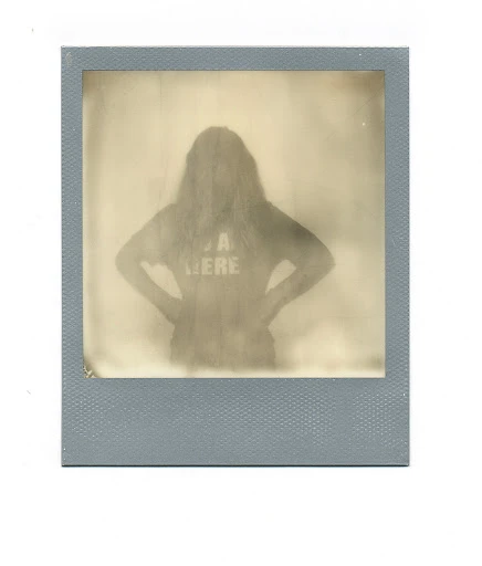 Monochrome photograph of a person with long hair wearing a t-shirt that reads, "I'M HERE", in a faded, square frame.