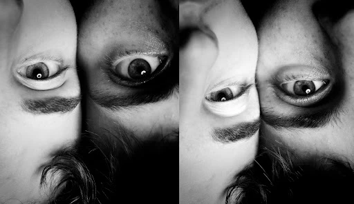 A black and white photograph featuring two faces in very close proximity to one another, eyes open and looking directly at the viewer.