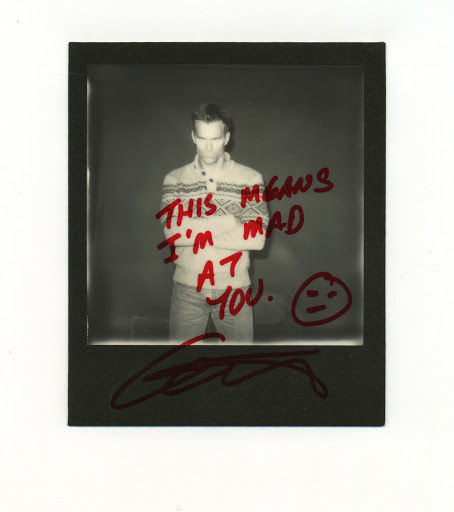Monochrome Polaroid of a man in a fair isle sweater with arms crossed, featuring hand-written text and signature on the print.