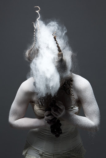 Artistic studio portrait of a female figure covered in white powder and a nude-toned garment against a neutral gray backdrop.