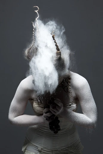 Artistic studio portrait of a female figure covered in white powder and a nude-toned garment against a neutral gray backdrop.