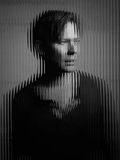 Monochrome portrait of an adult male with short, dark hair, featuring a striped overlay effect, set against a blurred background.