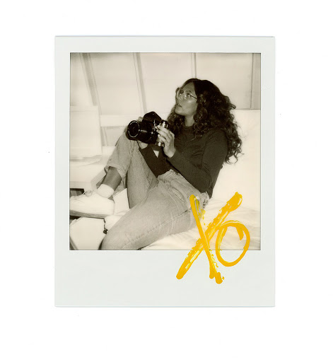 Polaroid photograph of a woman seated, holding a camera, with yellow 'XO' graffiti art in the foreground, indoor setting, light colour grading.