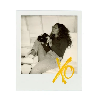 Polaroid photograph of a woman seated, holding a camera, with yellow 'XO' graffiti art in the foreground, indoor setting, light colour grading.