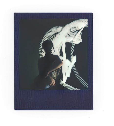 Polaroid portrait of a person with shoulder-length hair, wearing large earrings, against a dark background with light painting.
