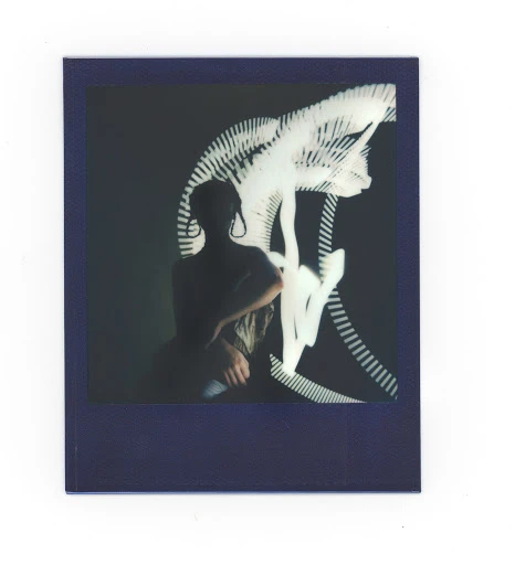 Polaroid portrait of a person with shoulder-length hair, wearing large earrings, against a dark background with light painting.