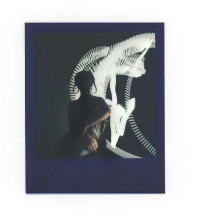 Polaroid portrait of a person with shoulder-length hair, wearing large earrings, against a dark background with light painting.