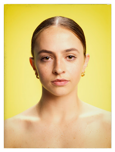 Close-up portrait of a young woman with brown hair and gold hoop earrings, against a bright yellow background in a studio shot.