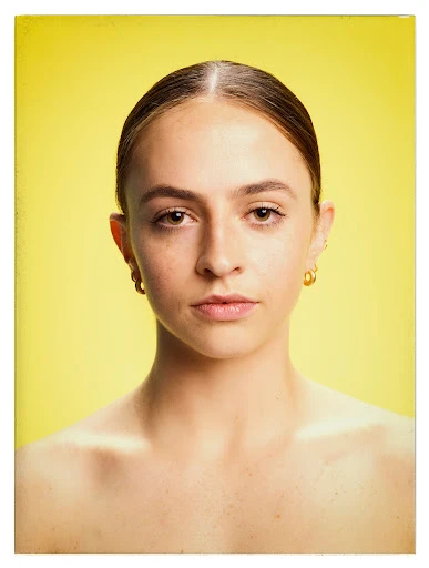 Close-up portrait of a young woman with brown hair and gold hoop earrings, against a bright yellow background in a studio shot.