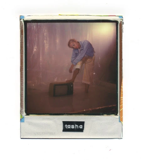 Woman in button-down shirt and striped pants poses with foot on small TV in retro-style photograph with textured border and name 'tasha'.