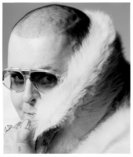 Black and white portrait of a person with a shaved head and sunglasses, wearing a fur-trimmed jacket, looking to the left.