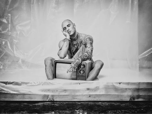 Black and white portrait of a bald, tattooed man sitting cross-legged on a vintage television, with a draped background, in a studio setting.