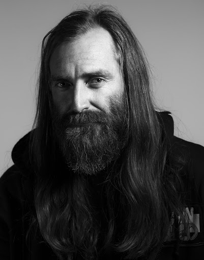 Black and white portrait of a bearded adult male with long hair wearing a dark hoodie against a neutral background.