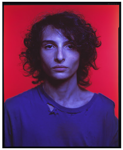 Portrait of a young adult with curly brown hair in a blue t-shirt against a vibrant red backdrop, bathed in a cool, blue light.